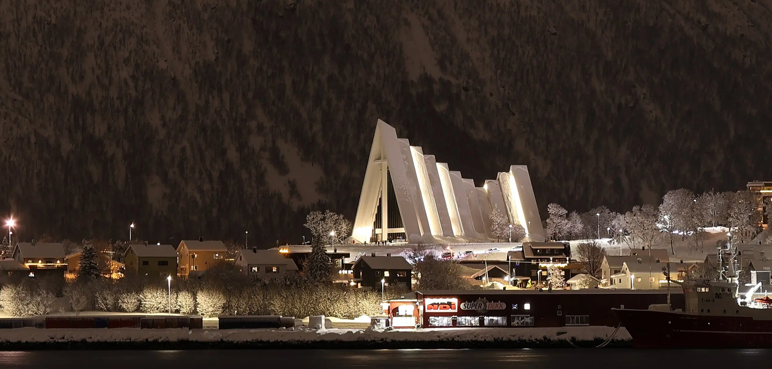Nighttime view of the illuminated Arctic Cathedral and surrounding snowy buildings in Tromsø