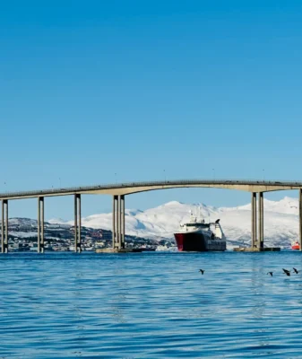 Big red fishing vessel passing under the Tromsø bridge on clear sunny day