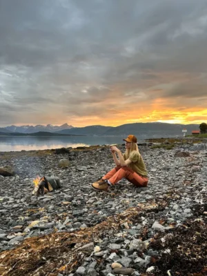 Person sitting by a small campfire on a rocky Arctic beach during golden sunset in Tromsø