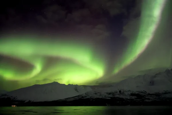 Bright spiral of Northern Lights glowing above snowy mountains and fjord near Tromsø
