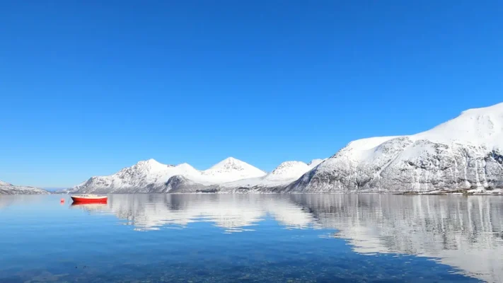 Red fishing boat anchored on calm crystal clear Arctic water near Sommarøy