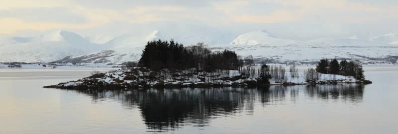 Small island with trees reflected in calm Arctic fjord waters against snowy mountains near Tromsø
