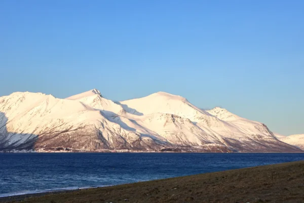 Snowy Arctic mountain range glowing in sunlight above deep blue fjord near Tromsø under clear sky
