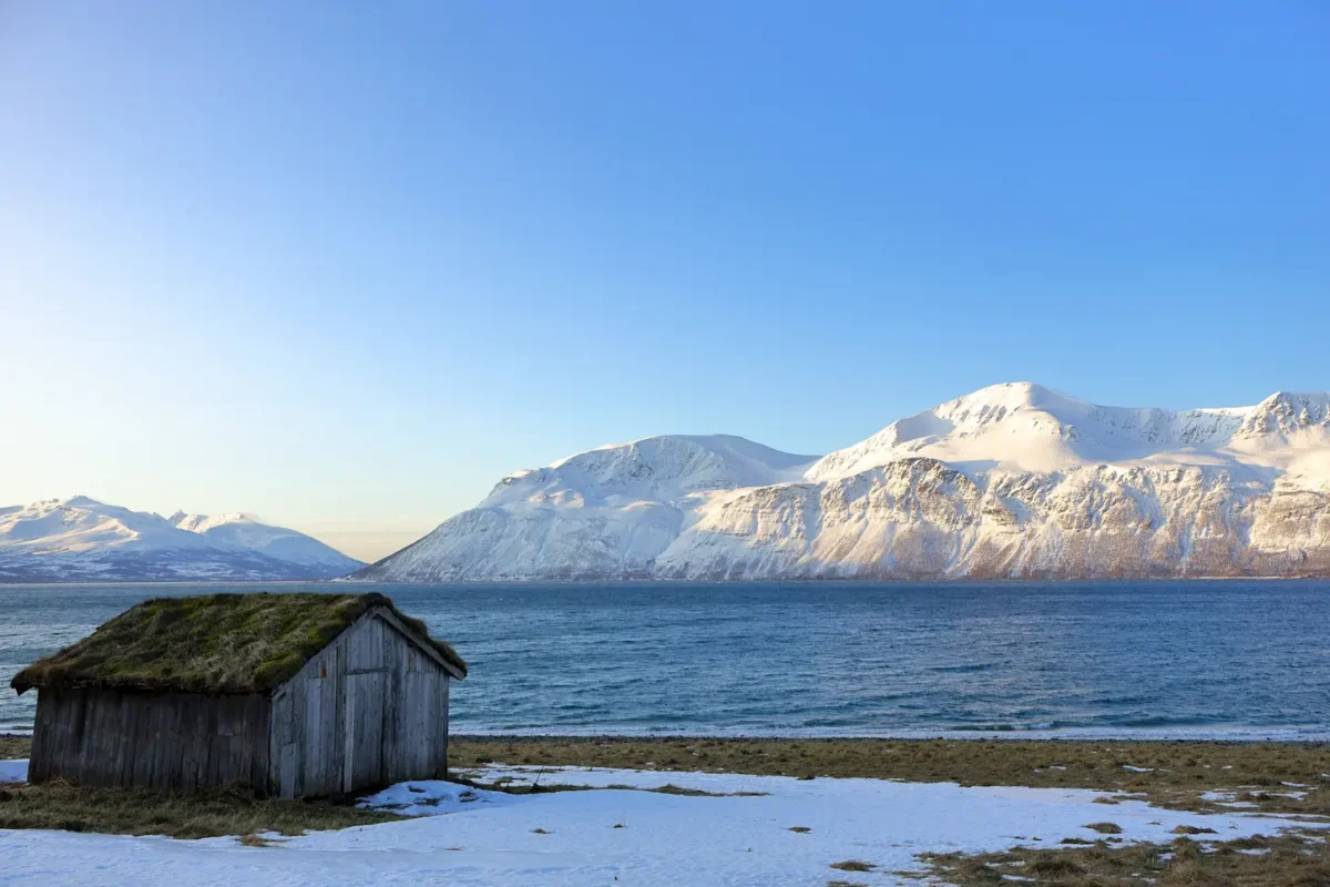 Old wooden hut with grass roof by Arctic fjord and sunlit snowy mountains near Tromsø