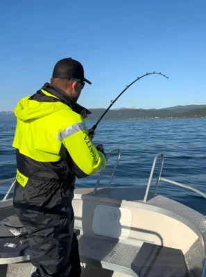 Man in a yellow and black fishing suit holding a bent fishing rod on a boat in Tromsø, reeling in a catch against a backdrop of calm Arctic waters and mountains.