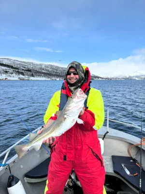 Smiling man in a red and yellow survival suit holding a freshly caught cod on a fishing boat in Tromsø, with snowy mountains and fjords in the background.
