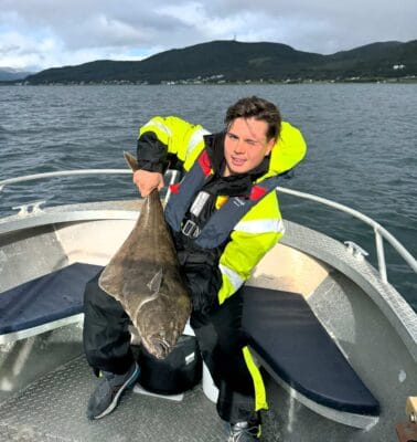 Man in a yellow and black fishing suit holding a large halibut on a boat during a fishing tour in Tromsø, with mountains and fjord in the background.