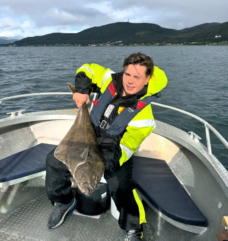 Man in a yellow and black fishing suit holding a large halibut on a boat during a fishing tour in Tromsø, with mountains and fjord in the background.