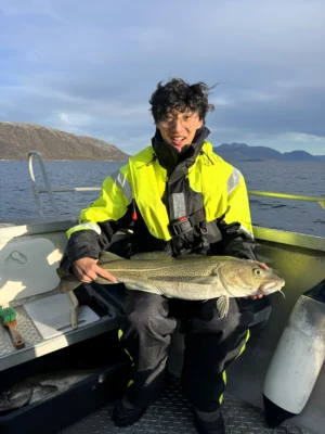 Smiling person holding a large cod on a boat during a private Arctic fishing tour near Tromsø, with mountain views and calm sea under clear skies.