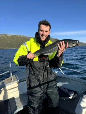 Person smiling while holding a saithe caught during a private Arctic fishing tour near Tromsø, under clear blue sky with mountain views.