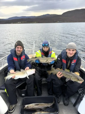 Three people sitting on a boat holding freshly caught cod during a private fishing tour near Tromsø, with calm fjord waters and mountains in the background.