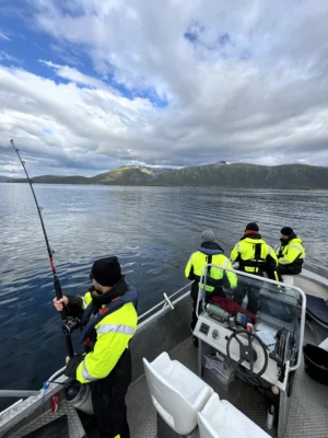 Group of people fishing from a boat in calm Arctic waters near Tromsø, surrounded by mountain views under a partly cloudy sky.