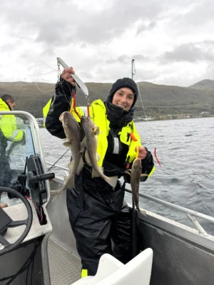 Smiling person on a boat holding several freshly caught cod during a private fishing tour near Tromsø, with fjord and mountain landscape in the background.